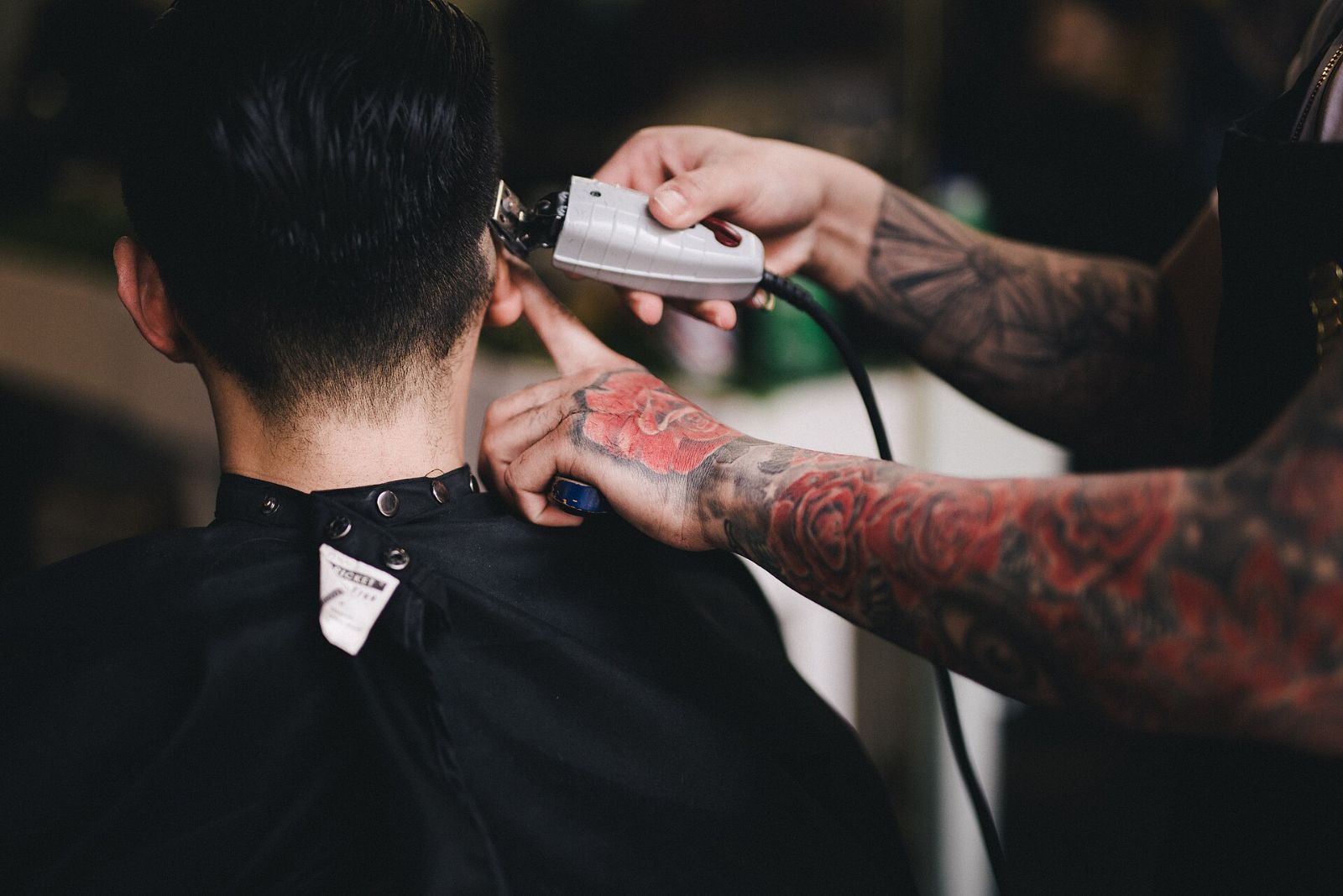 A barber with tattooed hands shaving the back of a customer's head with clippers