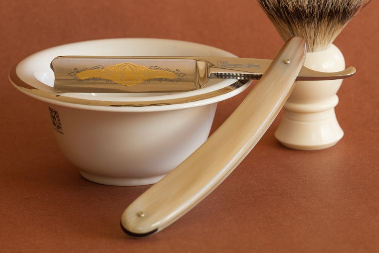 A straight razor, shaving brush and bowl on a brown background